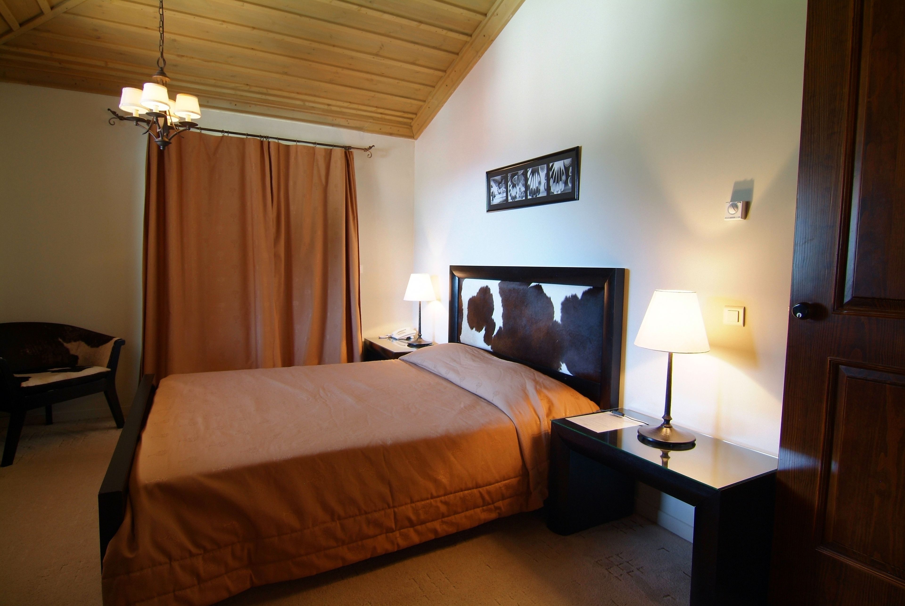 A hotel room with a bed, two bedside tables with lamps, a wooden headboard, a chair, and a wooden ceiling.
