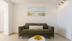 Una sala de estar con un sofá verde, una mesa de centro blanca con fruta, y una pintura de playa en la pared.