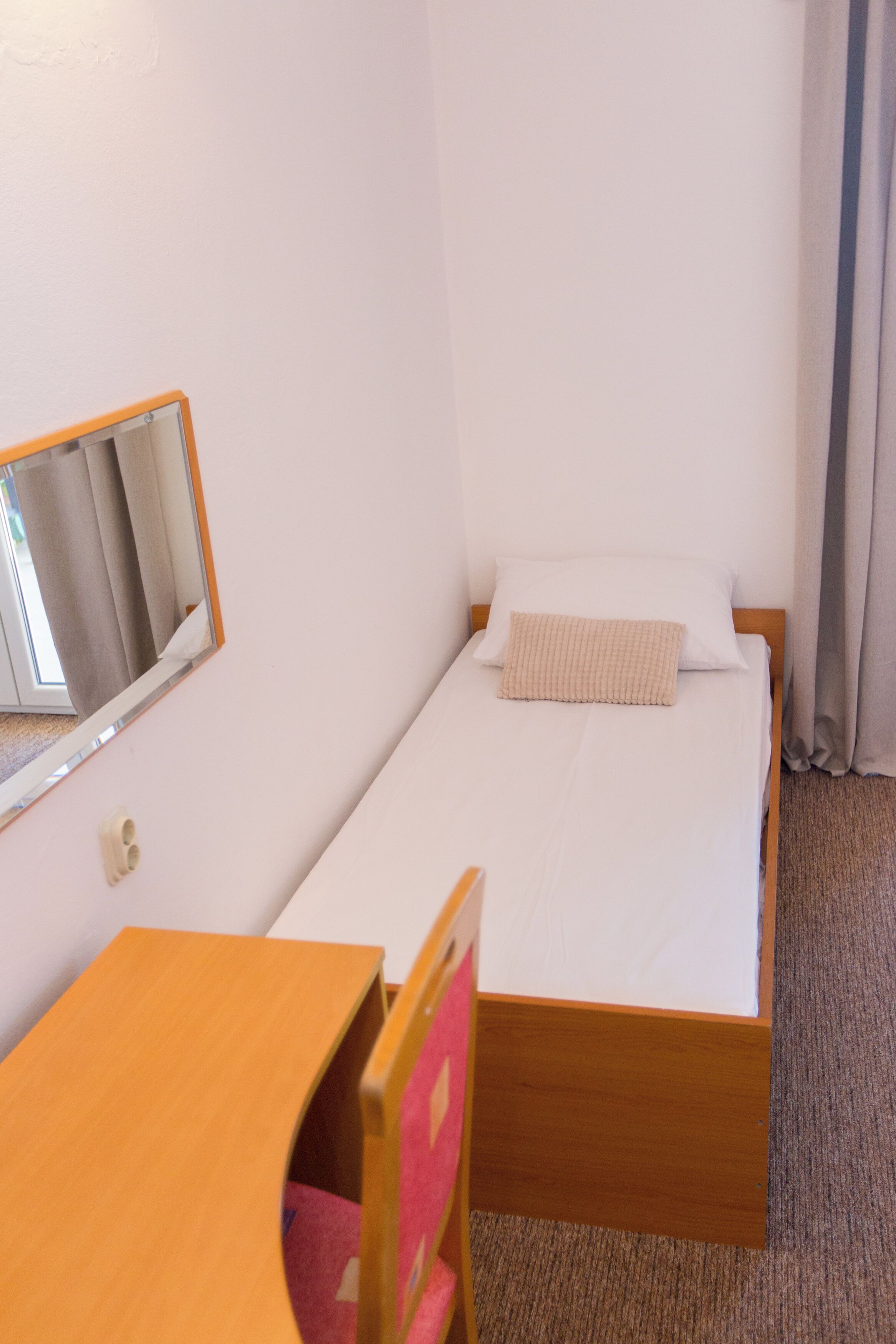 Une petite pièce simple, avec un lit simple, un bureau en bois, une chaise et un miroir au mur.