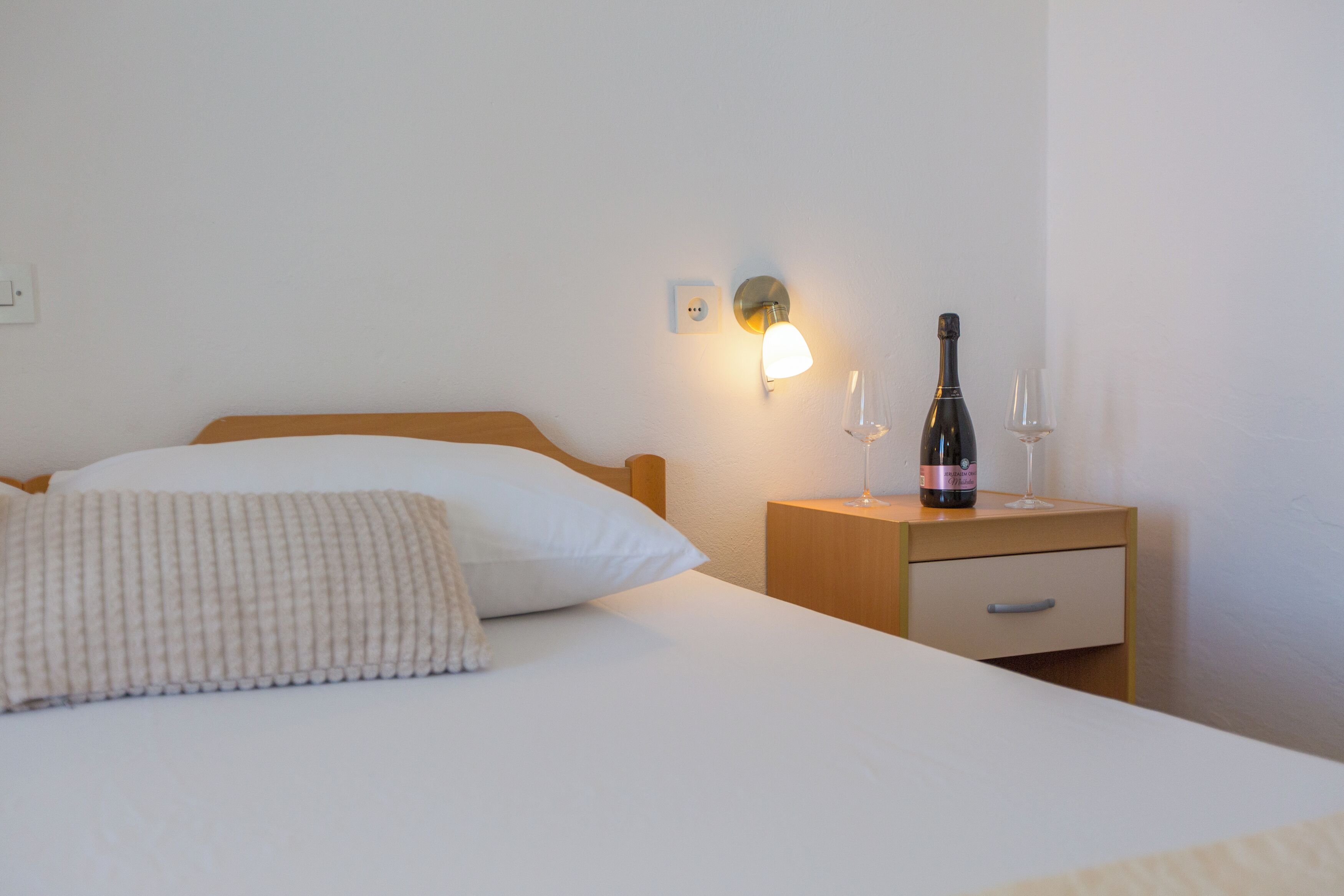 Ein ordentlich bezogenes Bett mit einem Kopfkissen und einem Nachttisch mit einer Weinflasche und zwei Gläsern.