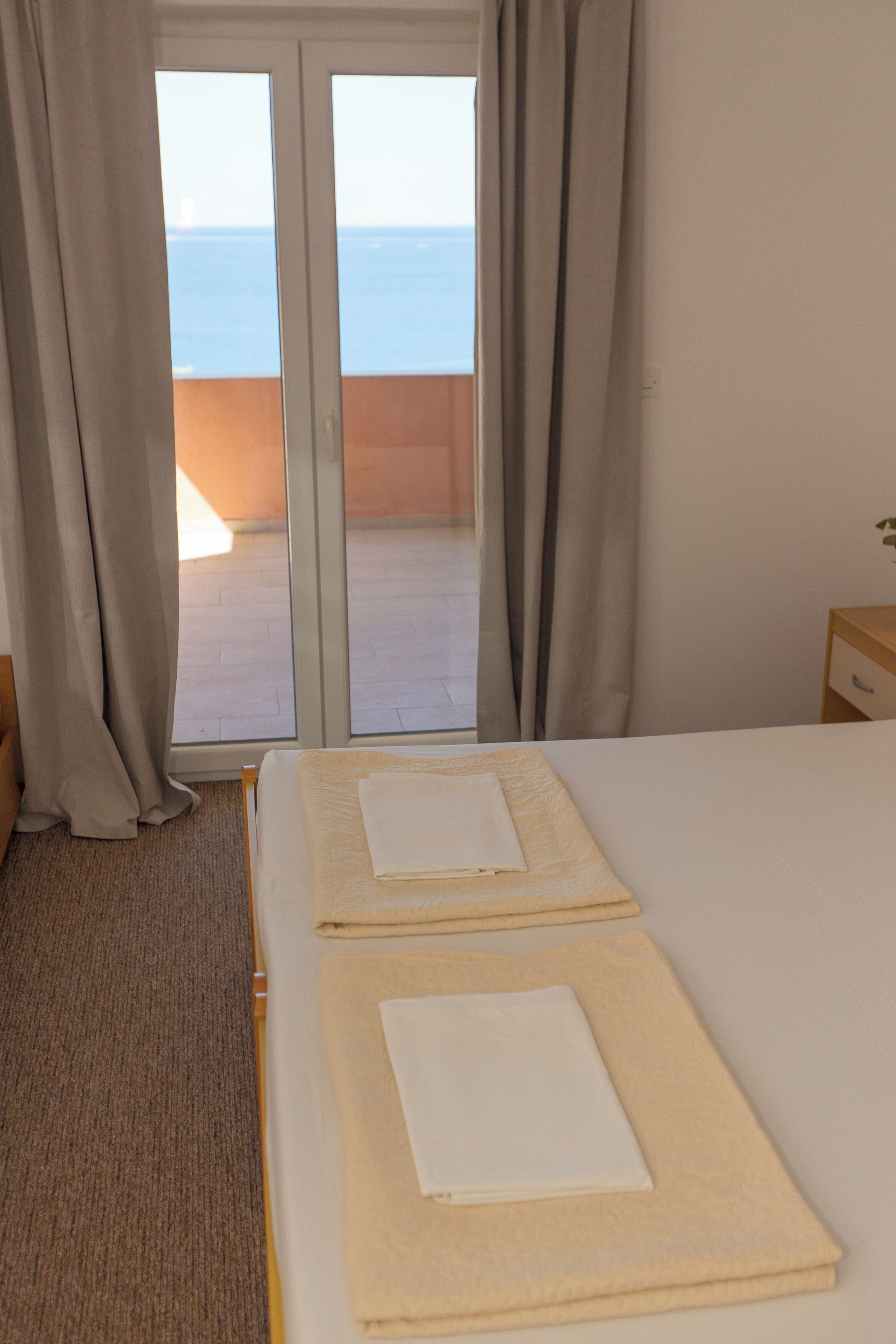 A hotel room with a bed, a wooden nightstand, and a view of the sea through a sliding glass door.