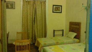 Um quarto pequeno com duas camas de solteiro, uma cadeira de madeira e uma janela com cortinas.