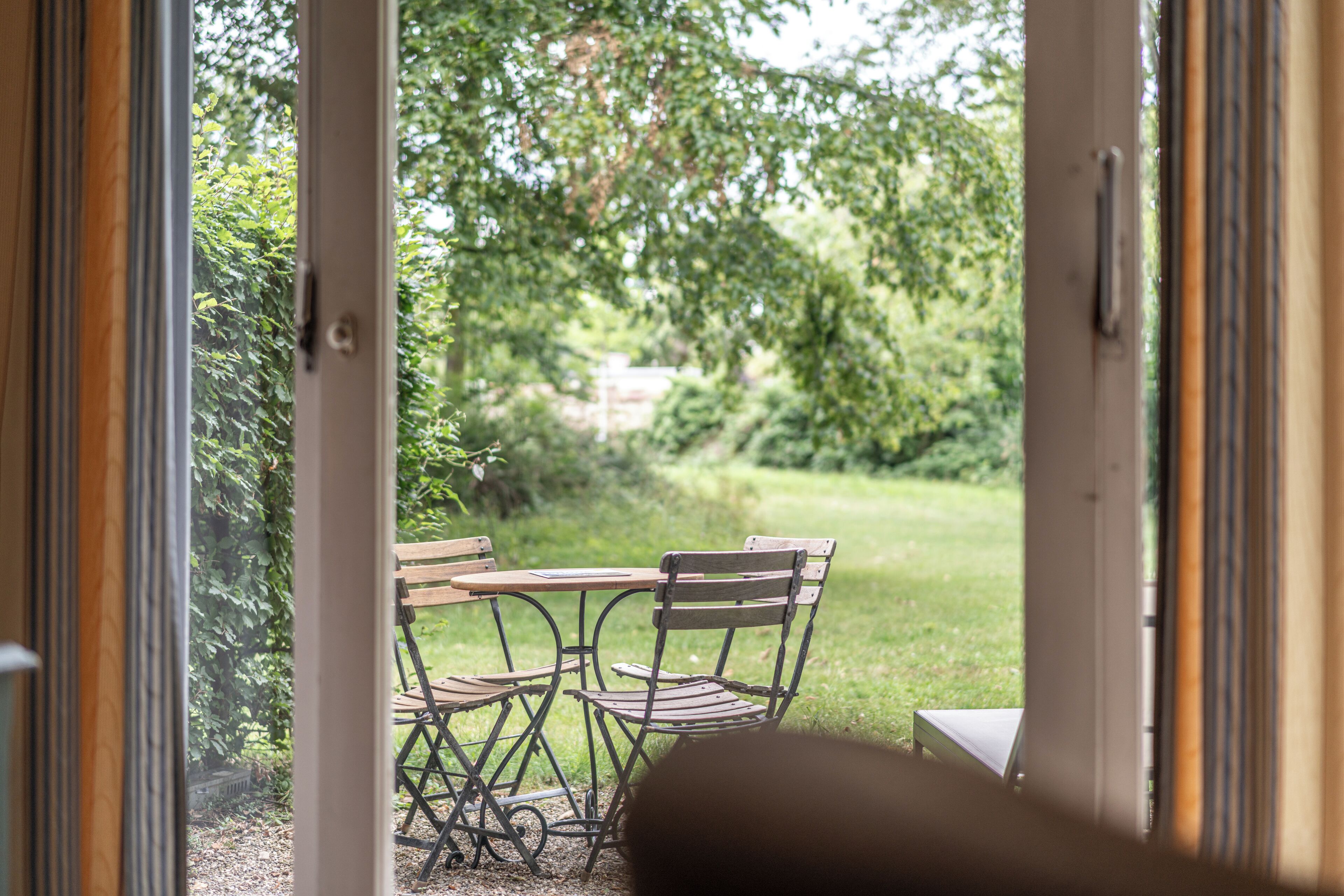 Een open deur die leidt naar een terras met een tafel en stoelen, omringd door groen.