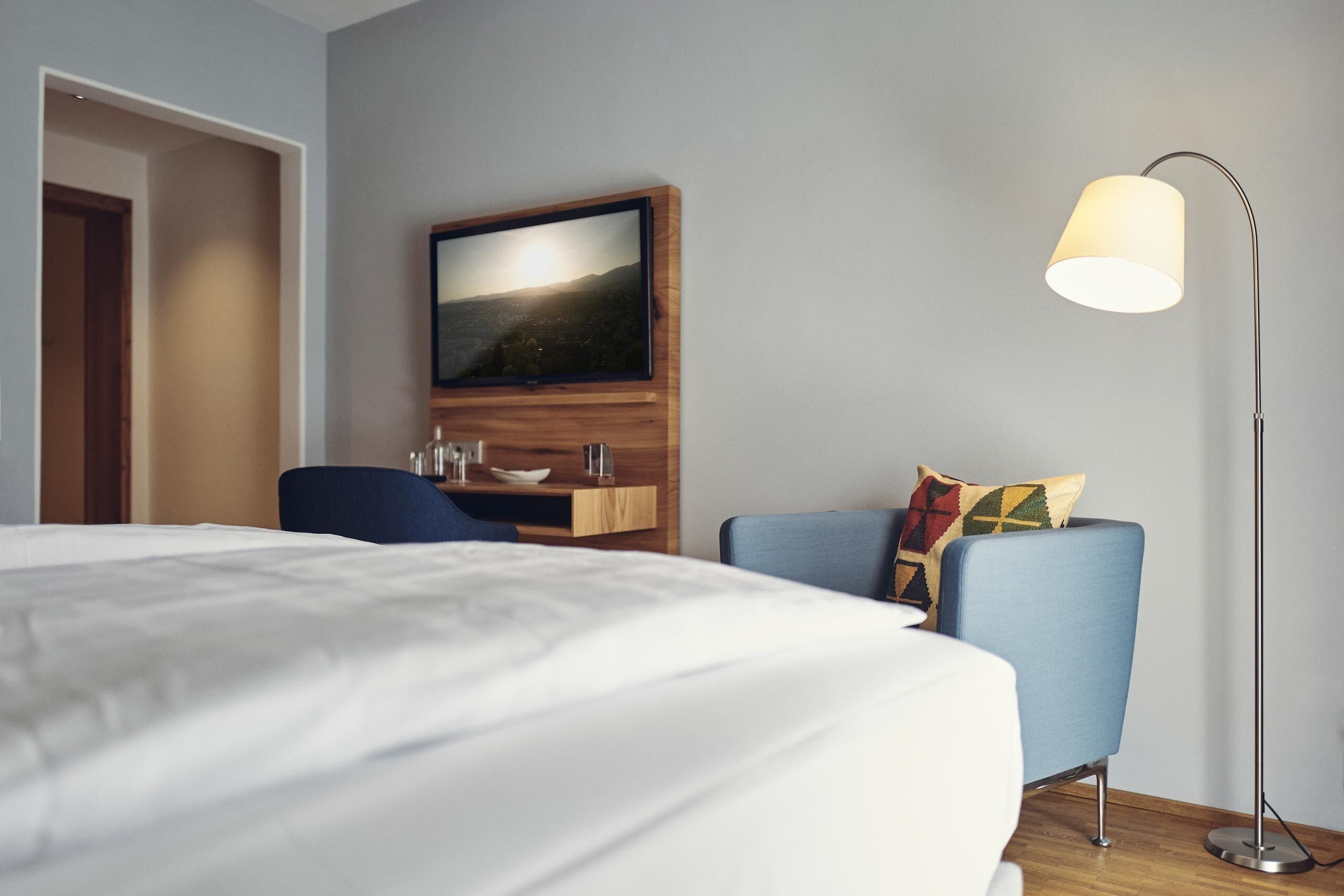 Ein Hotelzimmer mit Bett, Schreibtisch, Stuhl, Wand-Fernseher und Stehlampe.