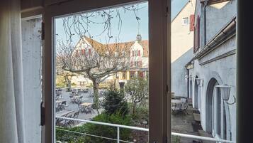 Vista desde un balcón con baranda metálica, sobre un patio con mesas y sillas al aire libre, un árbol y edificios con contraventanas rojas y una estructura con cúpula blanca.