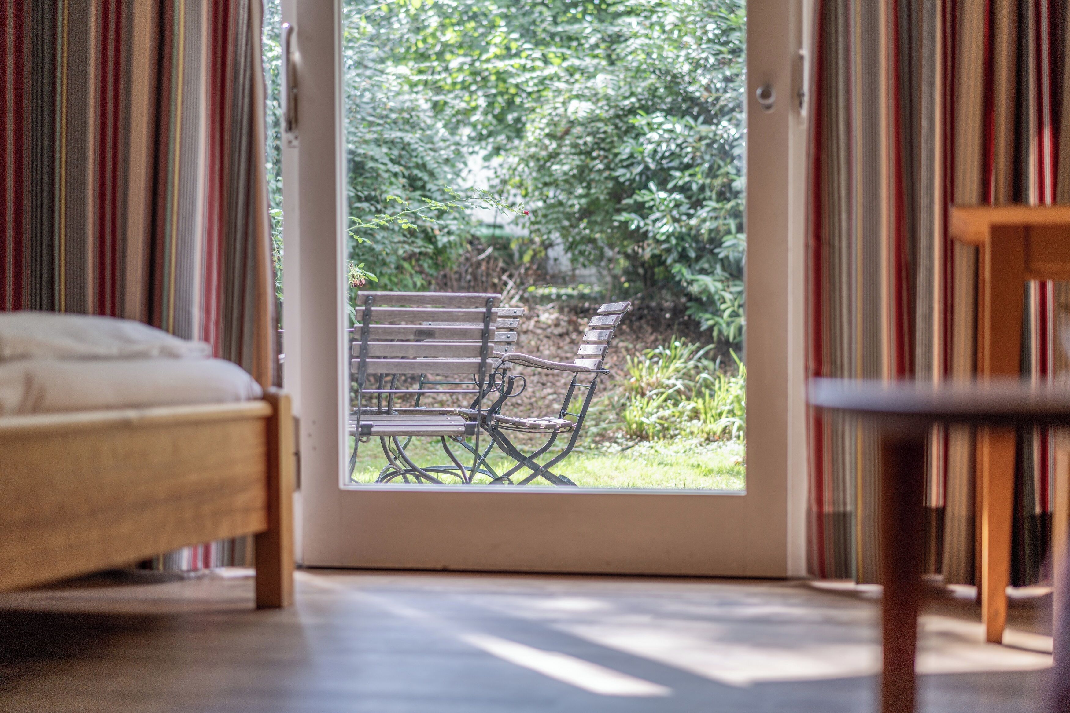 A room with a bed, a wooden table, and a bench outside a door leading to a garden.