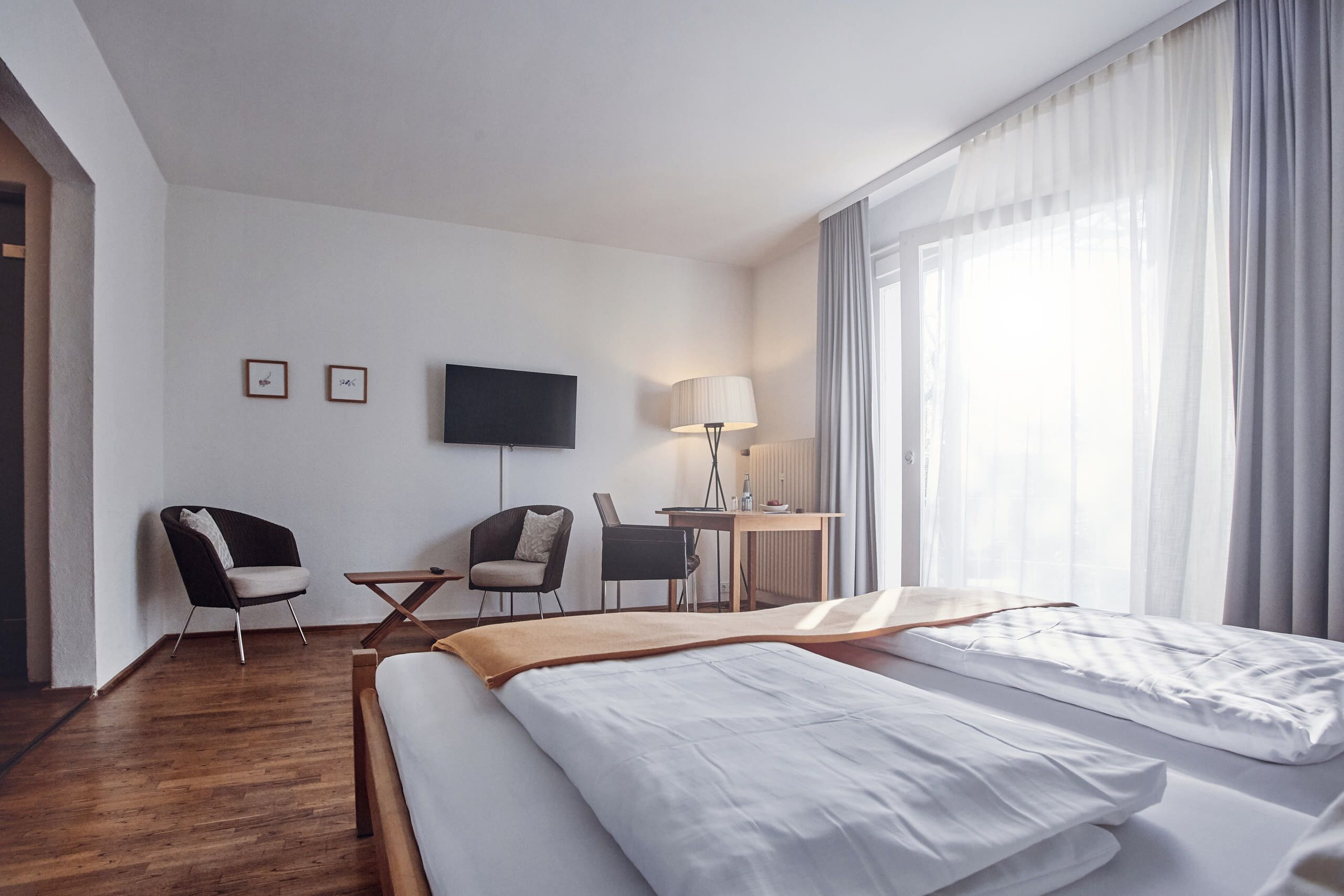 A hotel room with a bed, two chairs, a desk, a lamp, and a TV on the wall.