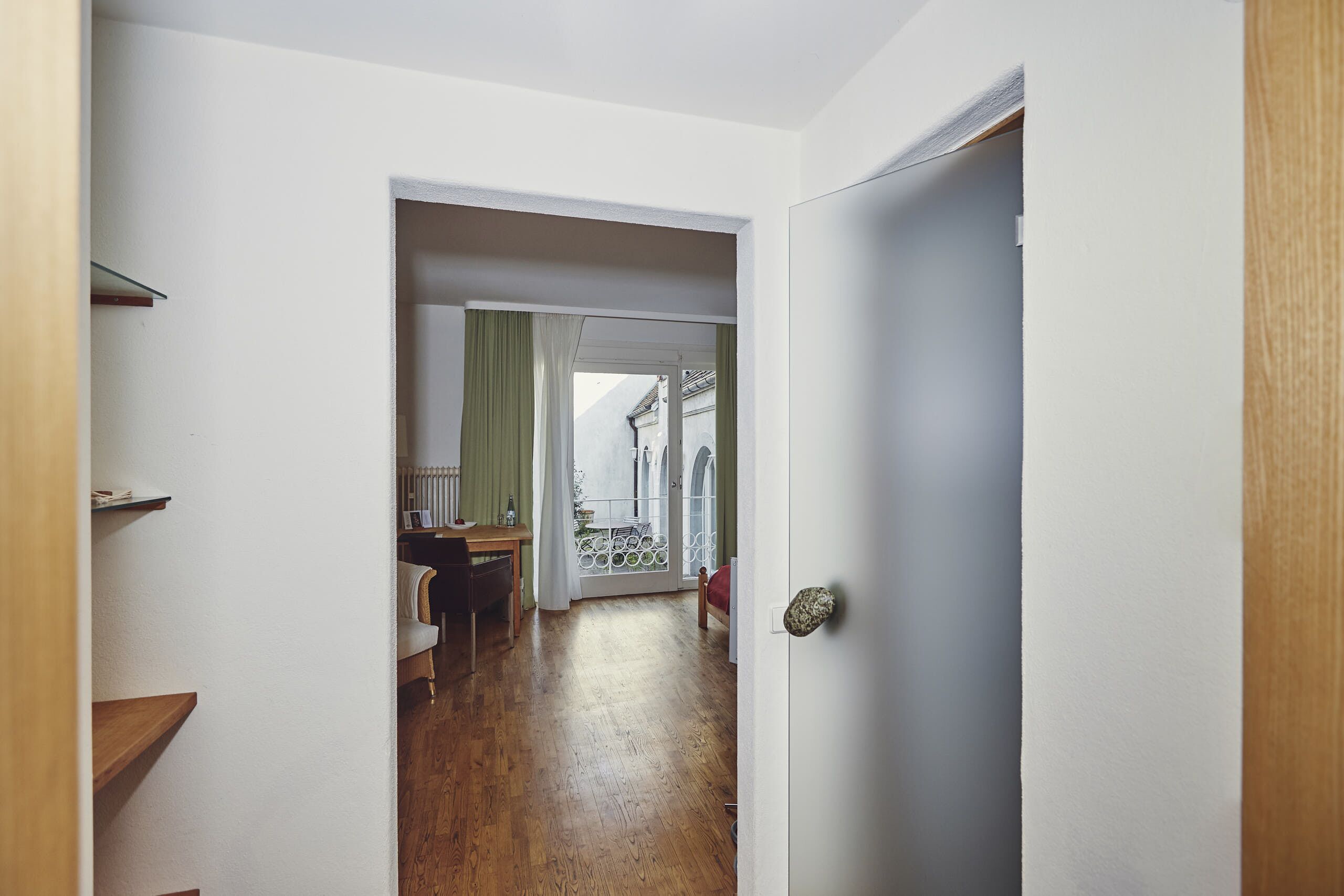 Un pasillo con piso de madera, una puerta a la derecha y vista a una habitación con una mesa y una silla.