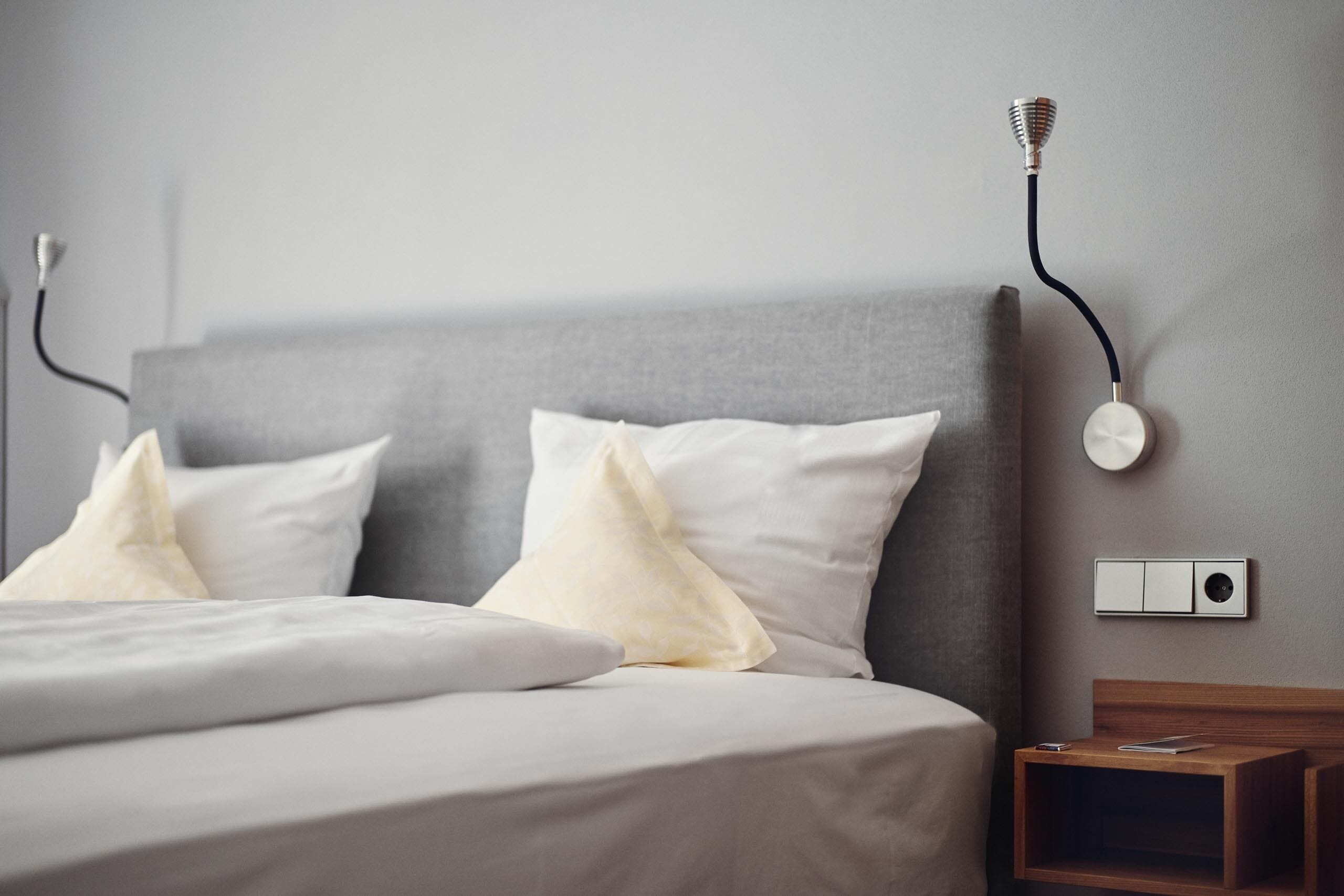 Cama bem arrumada com lençóis brancos e duas almofadas, mesa de cabeceira com abajur e luminária na parede.