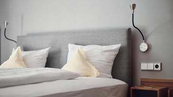 Cama bem arrumada com lençóis brancos e duas almofadas, mesa de cabeceira com abajur e luminária na parede.