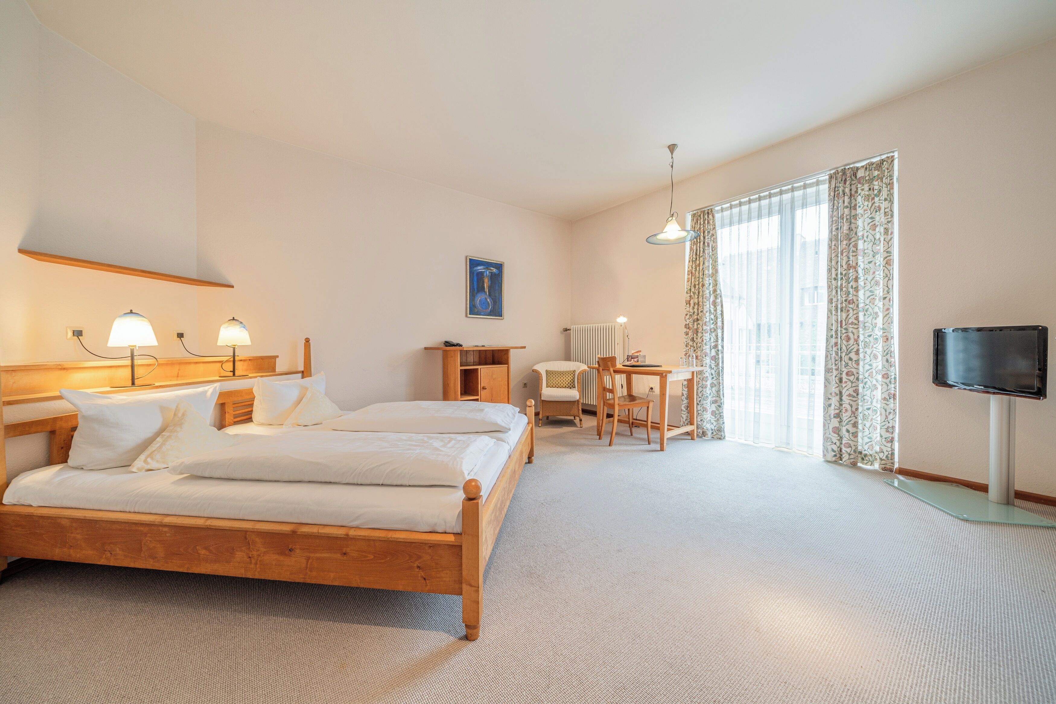 A hotel room with a wooden bed, a desk, a chair, a television, and a large window with curtains.