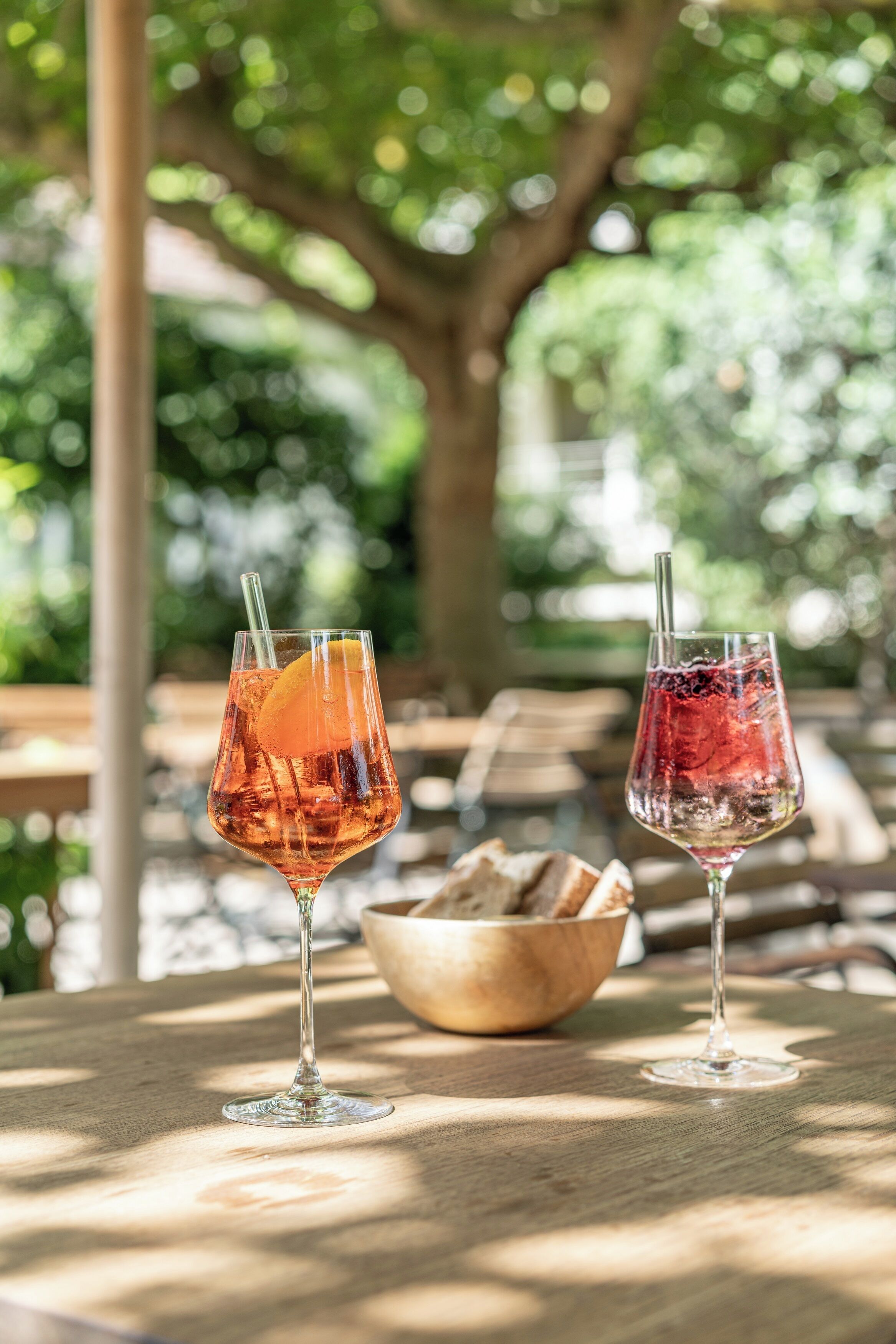 Dois copos de bebidas sobre uma mesa de madeira, com uma cestinha de pão ao fundo, em ambiente externo com árvores e uma estrutura de madeira.