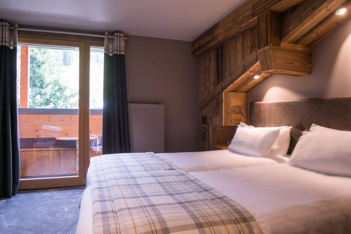 A bedroom with a large bed, wooden ceiling beams, and a view of an outdoor deck through a glass door.
