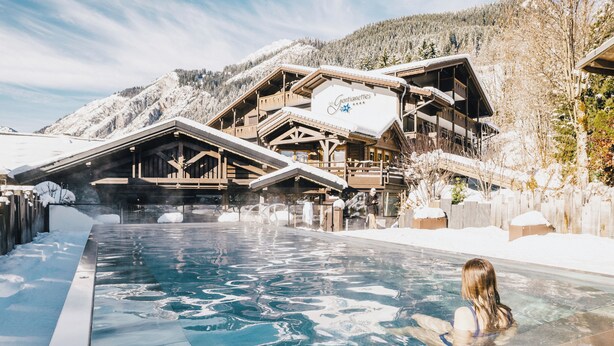 A pool area with a wooden building in the background, a person in the pool, and snow-covered mountains. - Chalet-Hôtel Les Gentianettes, The Originals Relais (Hôtels-Chalets de Tradition) (La Chapelle-d'Abondance)