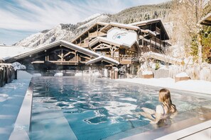 A pool area with a wooden building in the background, a person in the pool, and snow-covered mountains. - Chalet-Hôtel Les Gentianettes, The Originals Collection (Hôtels-Chalets de Tradition) (La Chapelle-d'Abondance)