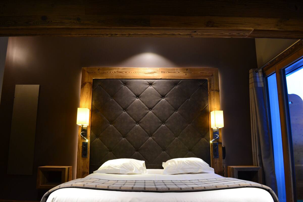 A hotel room with a tufted headboard, two bedside lamps, a bed with white linens, and a wooden ceiling.