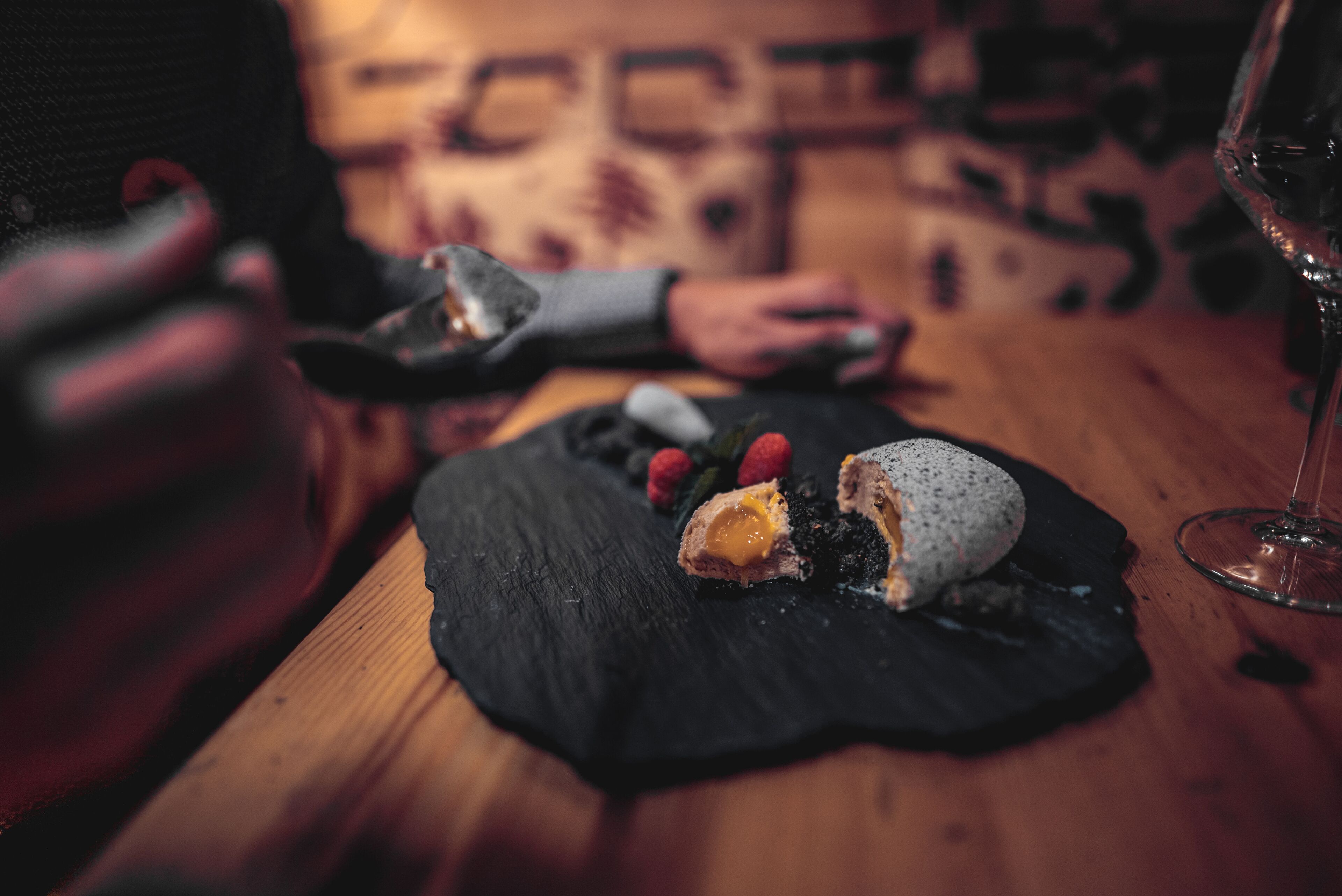 A dining table with a dessert plate, a glass of wine, and a person holding a phone.