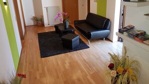 Un salon moderne avec un canapé noir, une table basse et une petite table d’appoint. On y trouve une bibliothèque remplie de livres et une plante en pot.