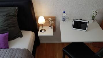 A hotel room with a bed, a bedside table with a lamp, a water bottle, a glass, a tablet, and a small potted plant.