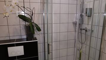 Un baño con ducha de vidrio, paredes de azulejos blancos, encimera negra y una orquídea en maceta.