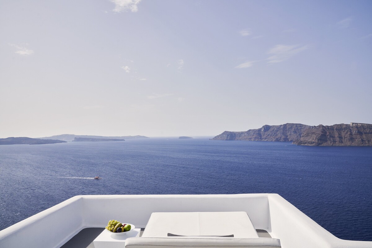 A boat deck with a white table, a bowl of fruit, and a view of the sea and distant cliffs.