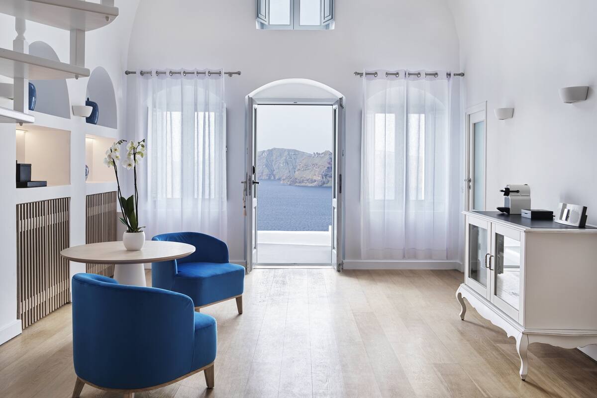 A modern room with a view of the sea, featuring a round table, blue chairs, a white cabinet, and a potted orchid.