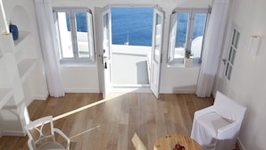 A room with a wooden floor, white walls, and a view of the sea through open doors. There is a white chair, a small round table with apples, and a potted plant.