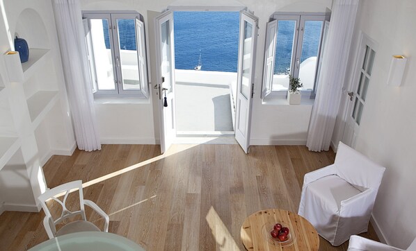 A room with a wooden floor, white walls, and a view of the sea through open doors. There is a white chair, a small round table with apples, and a potted plant. - Katikies Santorini - The Leading Hotels of the world (Santorini)