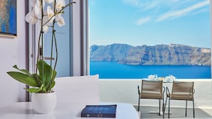 A room with a view of a mountainous coastline, a potted orchid, a book on the table, and two chairs. - Katikies Santorini – Pelagos House - The Leading Hotels of the World (Santorini)