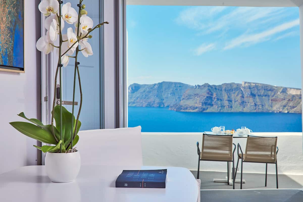 A room with a view of a mountainous coastline, a potted orchid, a book on the table, and two chairs.