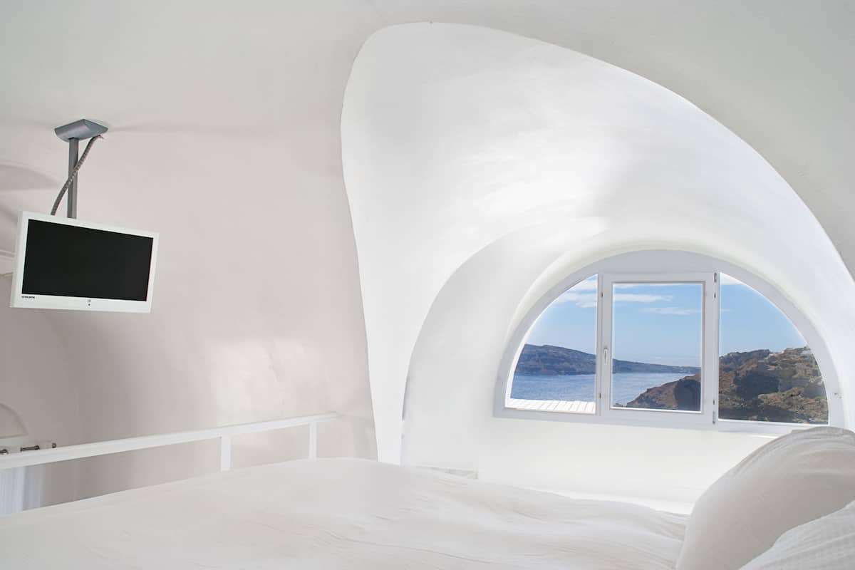 A bedroom with a curved ceiling, a bed, a TV mounted on the wall, and a window with a view of the sea.