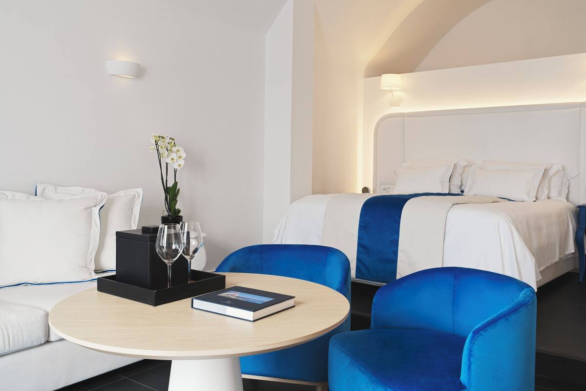 A hotel room with a bed, two blue armchairs, a round table with a book and a vase of flowers, and a white sofa.