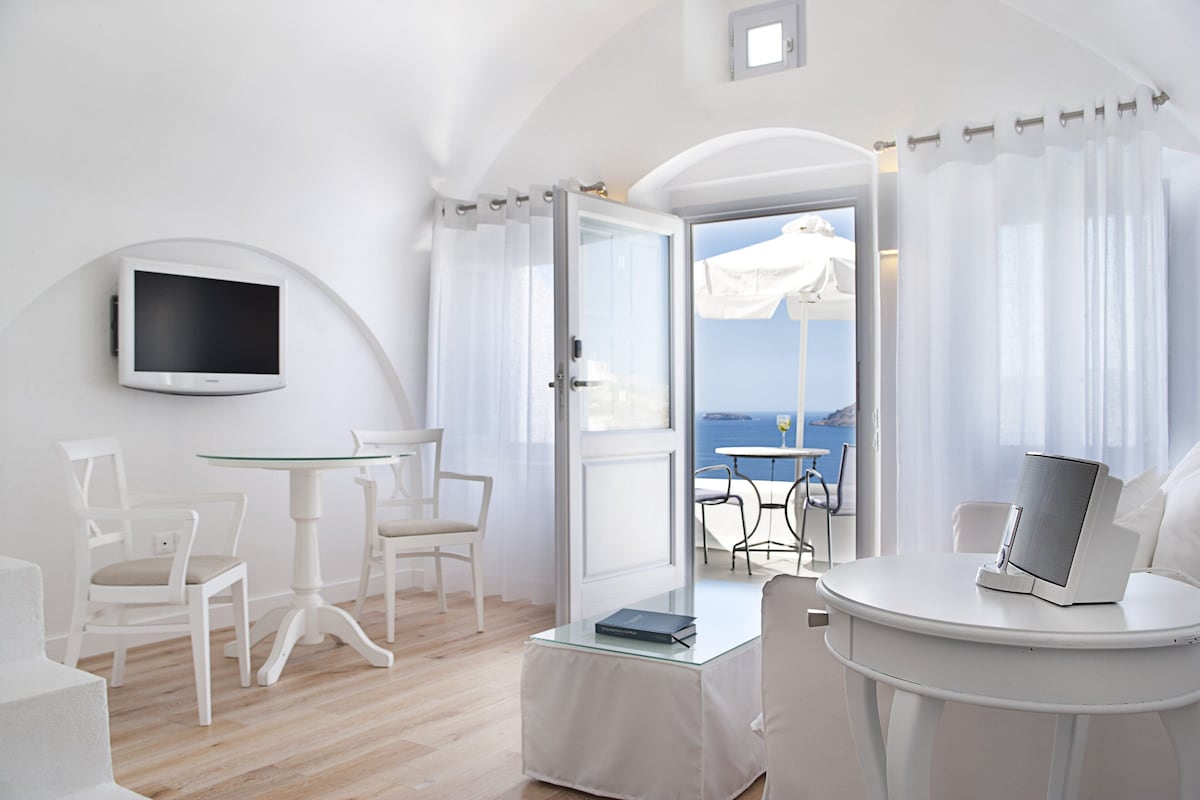 Una habitación moderna con decoración blanca, un televisor montado en la pared, una mesita redonda con un libro y vista al mar por una puerta abierta.