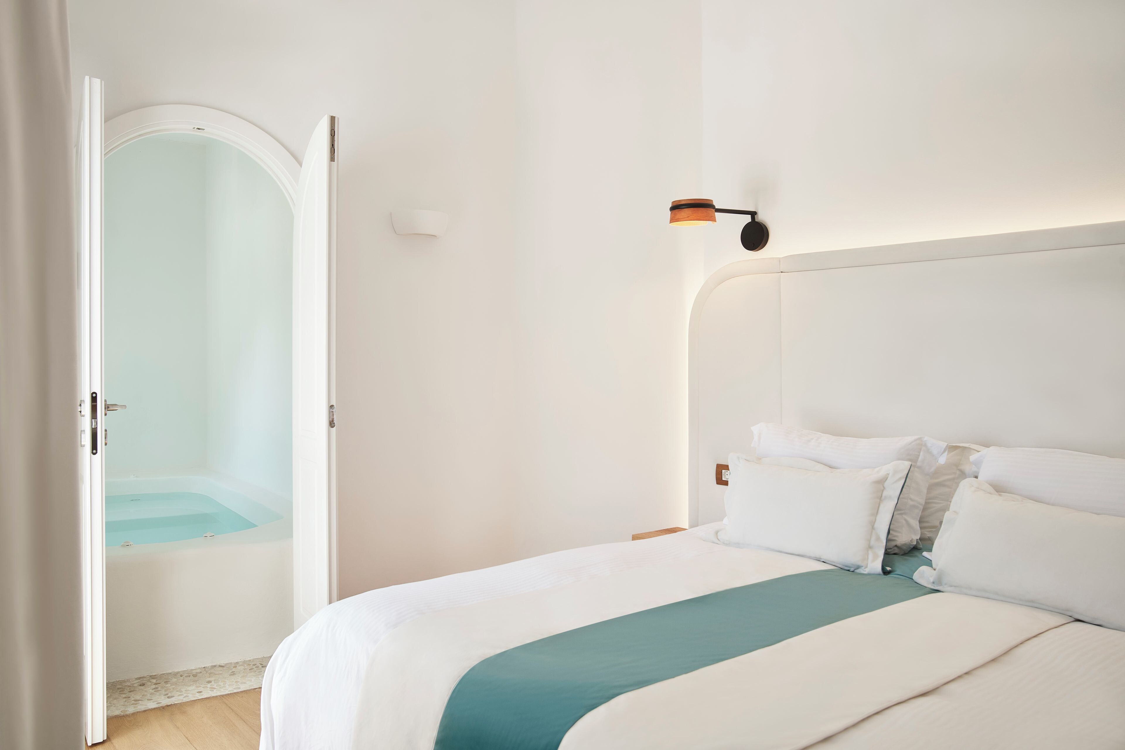 A hotel room with a bed, a nightstand, a wall-mounted lamp, and an open door leading to a bathroom with a bathtub.