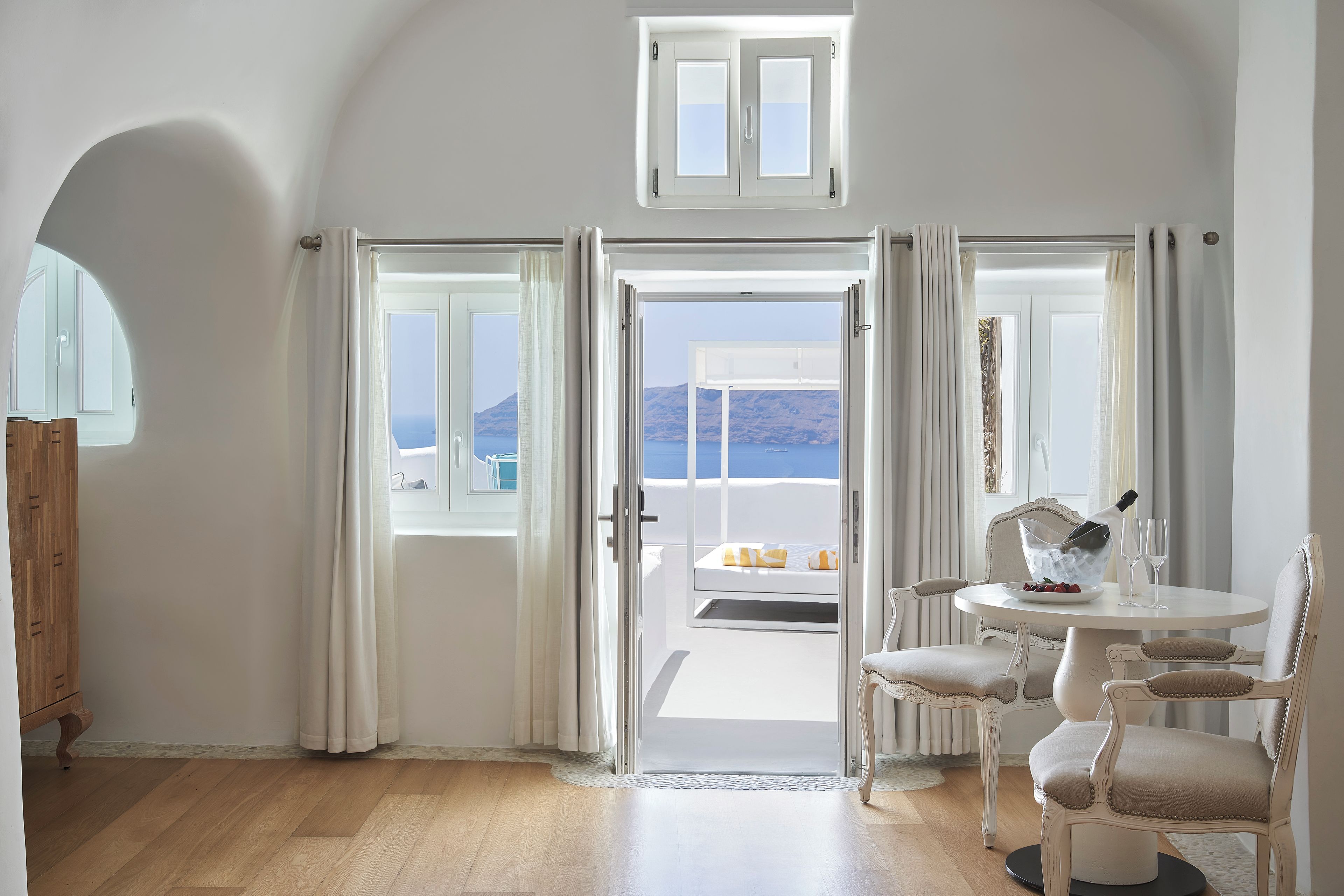 A room with a view of the sea, a balcony with a table and chairs, and a wooden floor.