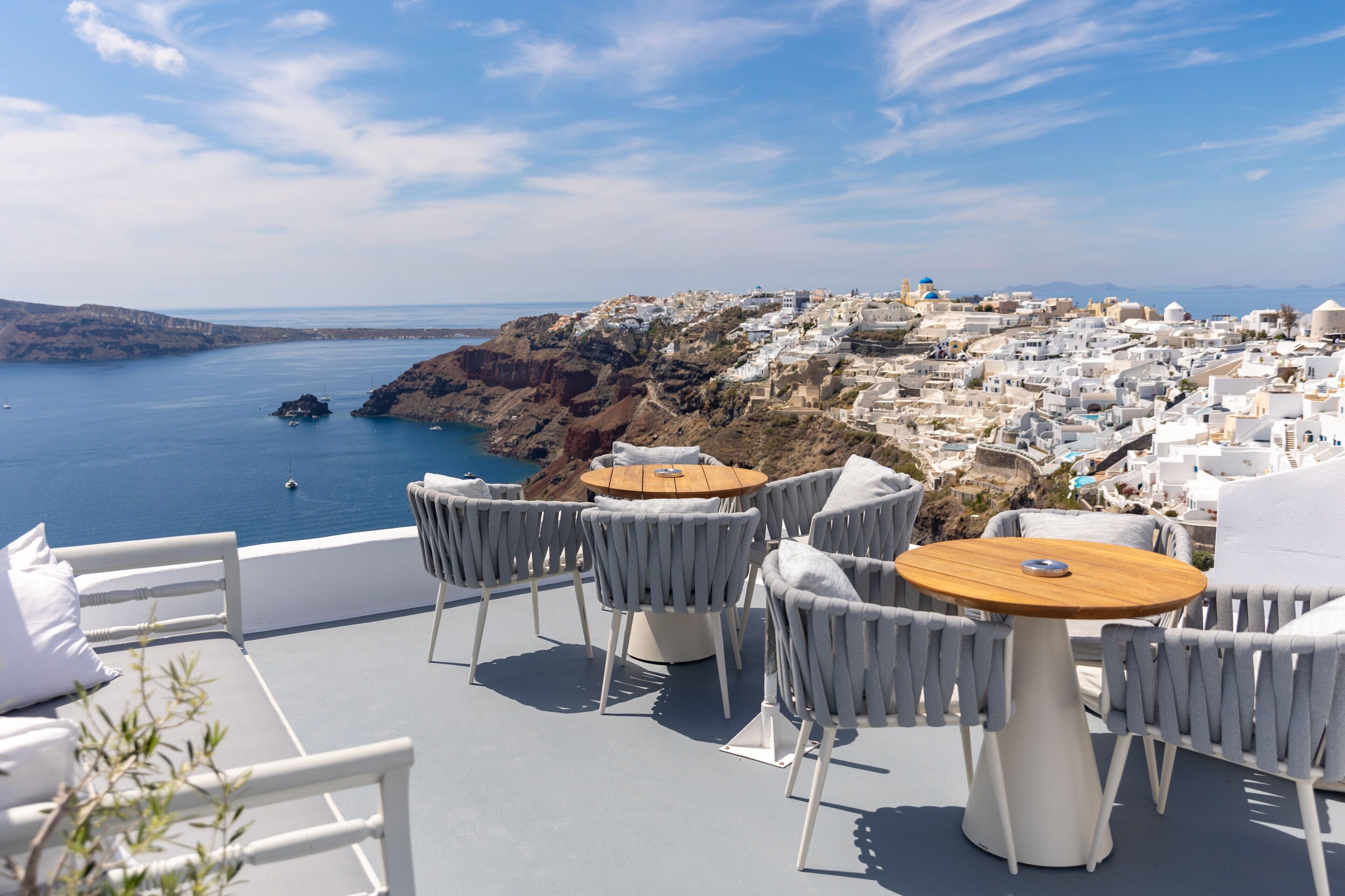 A terrace with white furniture overlooking a coastal town with white buildings and a view of the sea.
