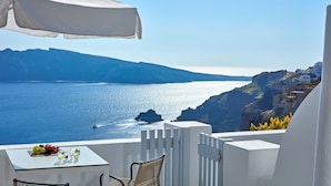 A balcony with a table, a chair, and a white umbrella overlooking a coastal view with a boat on the water and buildings on the cliff.