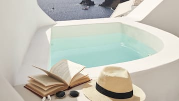 Una piscina infinita blanca con vista al mar y acantilados, un libro y un sombrero de paja sobre una superficie blanca.