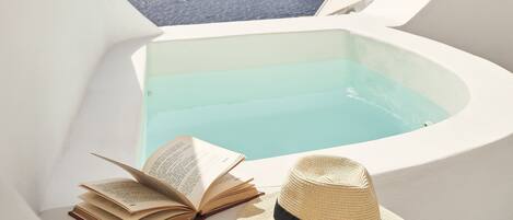 A white infinity pool with a view of the sea and cliffs, a book, and a straw hat on a white surface.