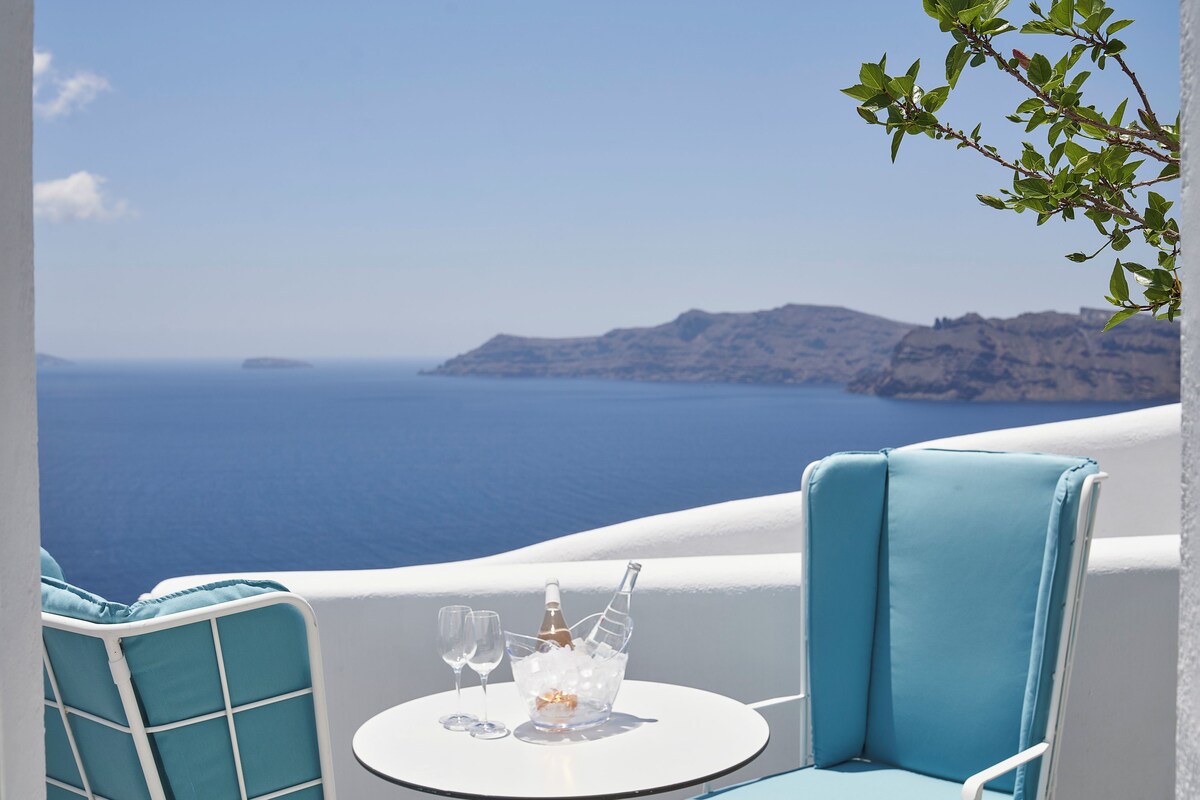 A balcony with a table set for two, a bottle of champagne, and two glasses. The view includes a body of water and distant landforms.