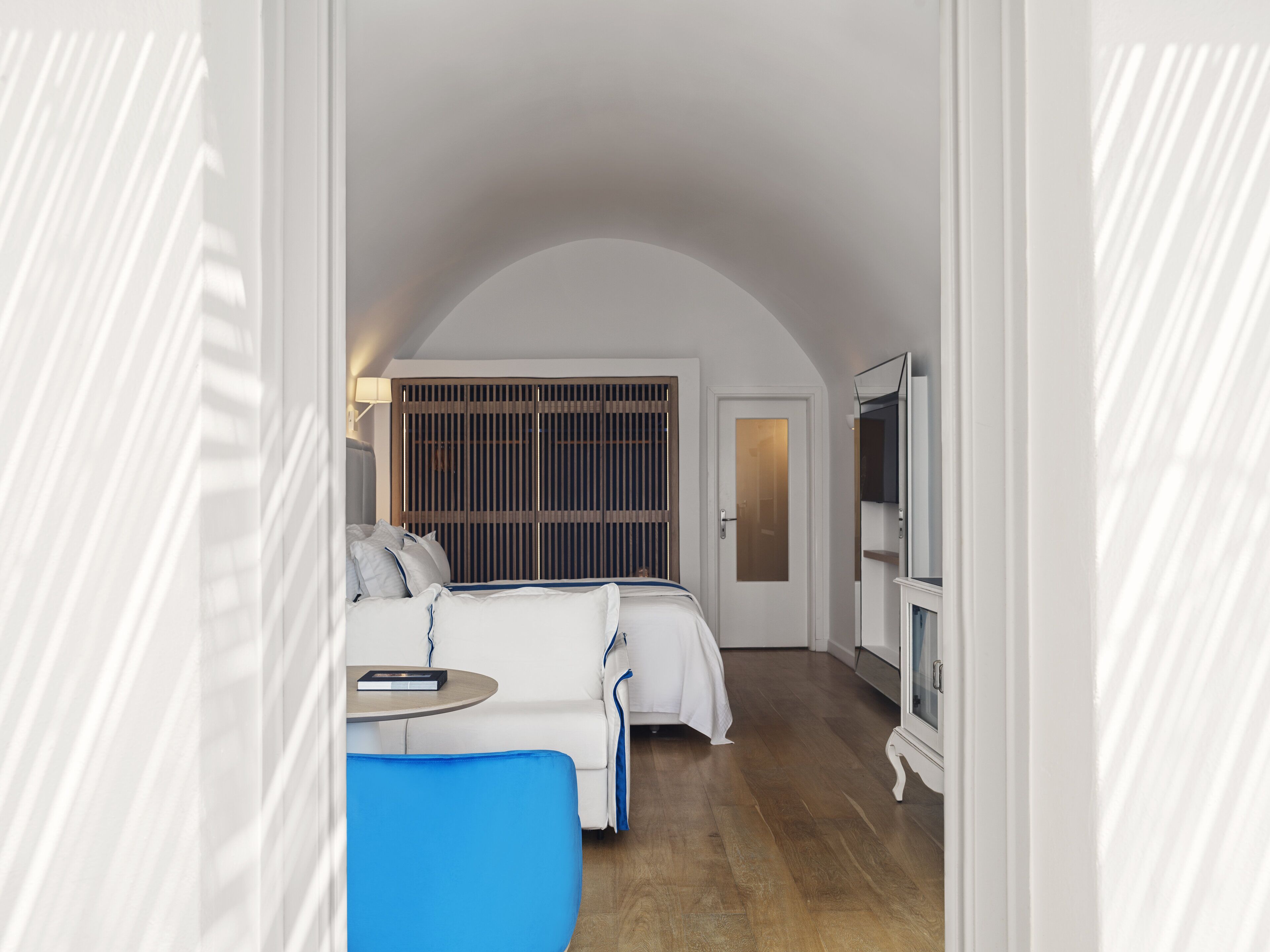 A hotel room with a bed, a small table, a blue chair, a TV, and a wardrobe.