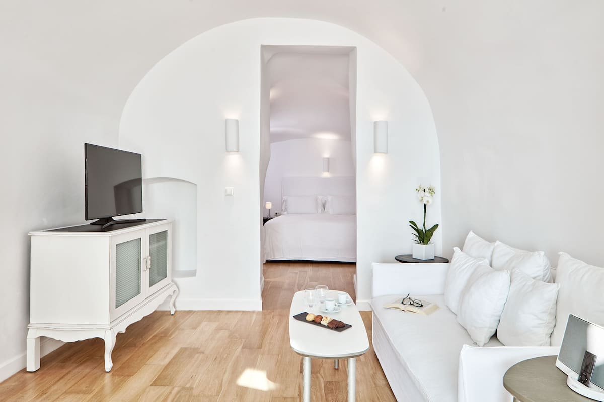 A modern hotel room with a white color scheme, a flat-screen TV, a small dining table, a sofa, and a bed with white bedding.