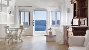 A spacious room with a large window offering a view of the sea and mountains. There is a dining table with chairs, a white sofa, and a wooden cabinet.