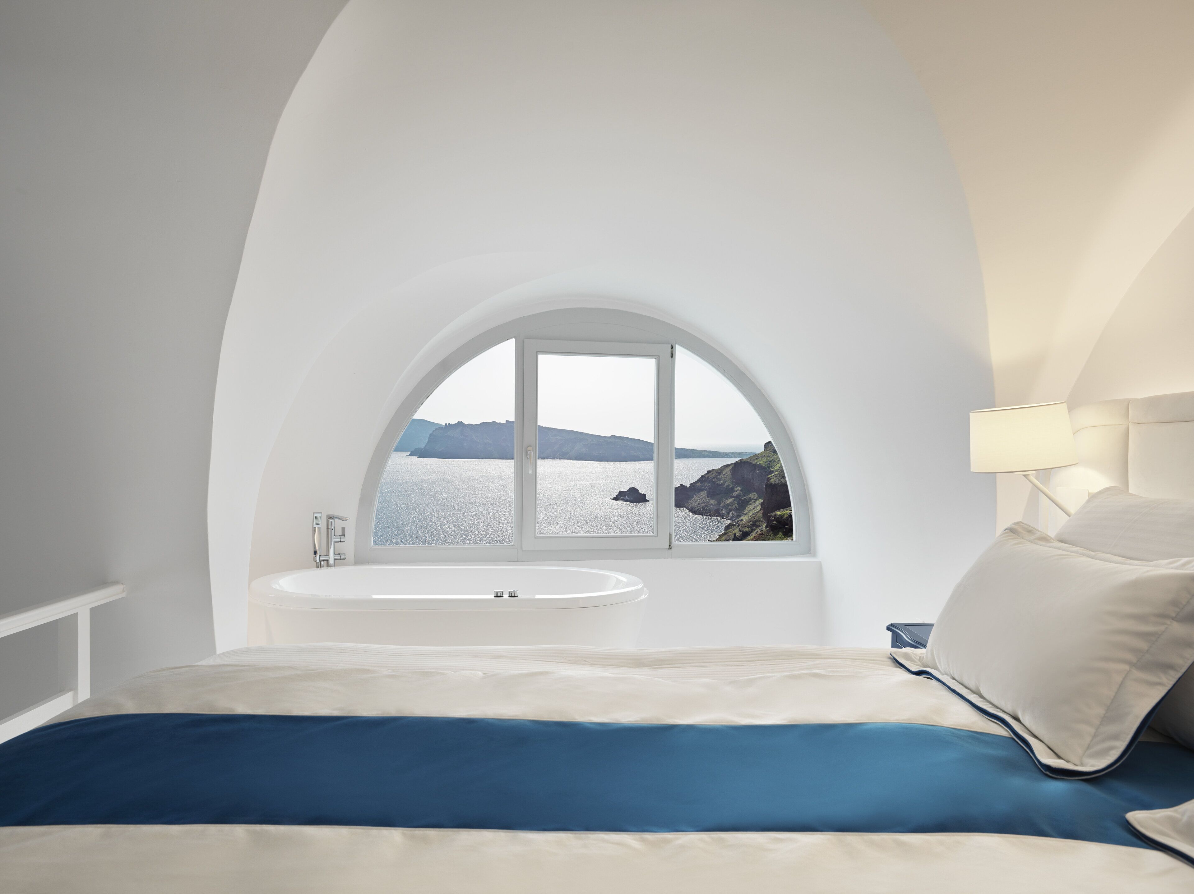 Un dormitorio con techo blanco curvo, un ventanal arqueado grande con vistas al mar y acantilados, una bañera blanca y una cama con ropa de cama azul y blanca.