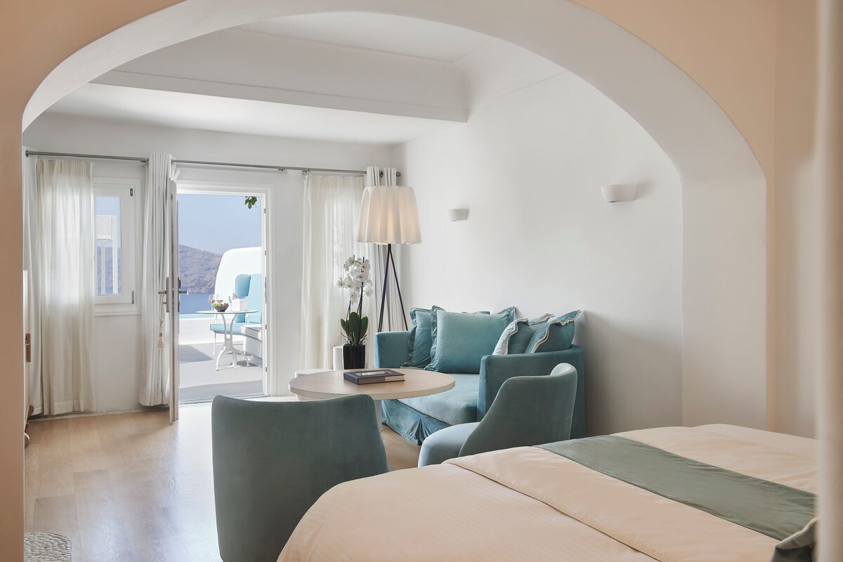 A modern hotel room with a bed, a sofa, a small table, and a view of the outdoors.