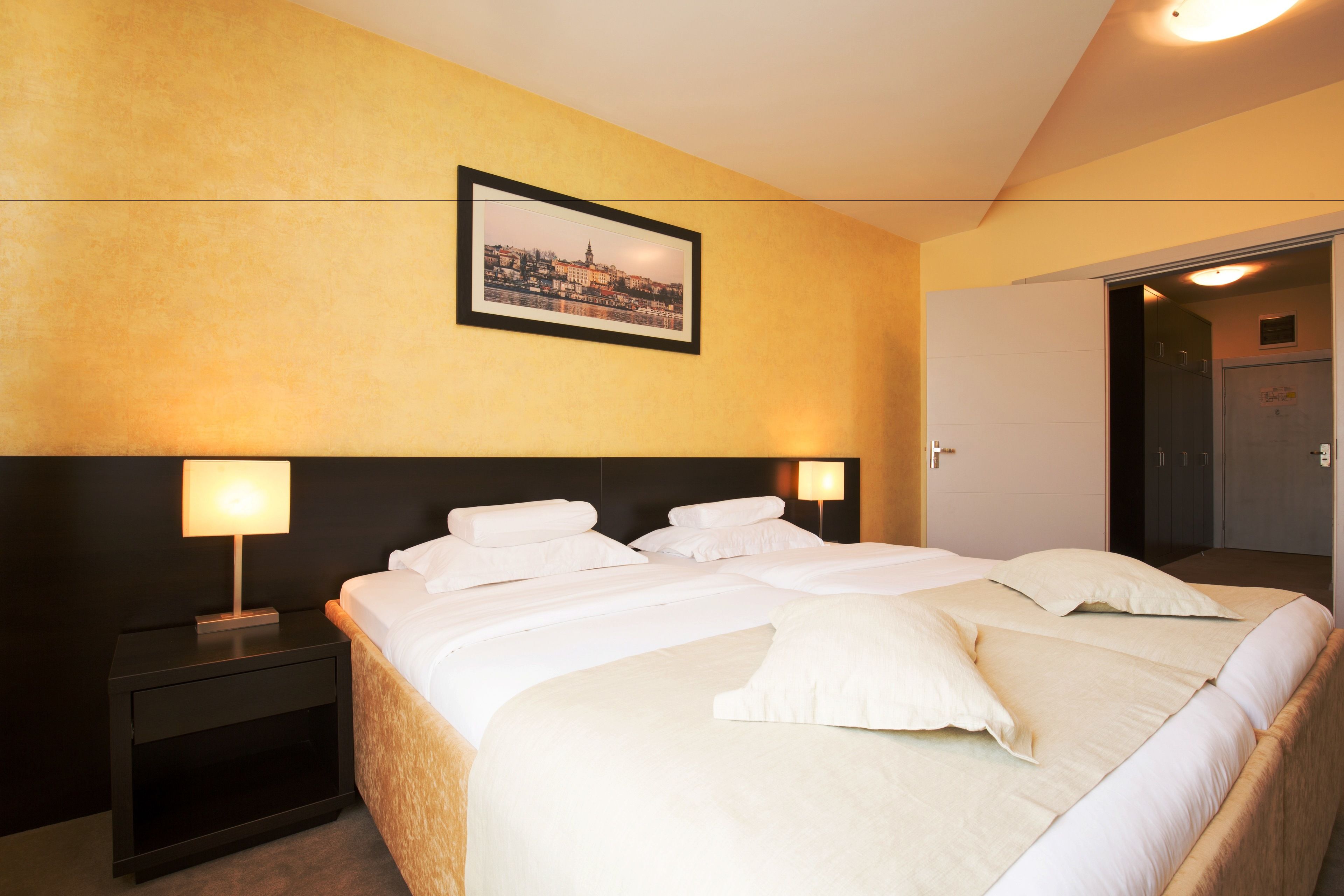 Quarto de hotel com uma cama grande, duas mesas de cabeceira com luminárias, um quadro na parede e uma porta que dá para outro quarto.