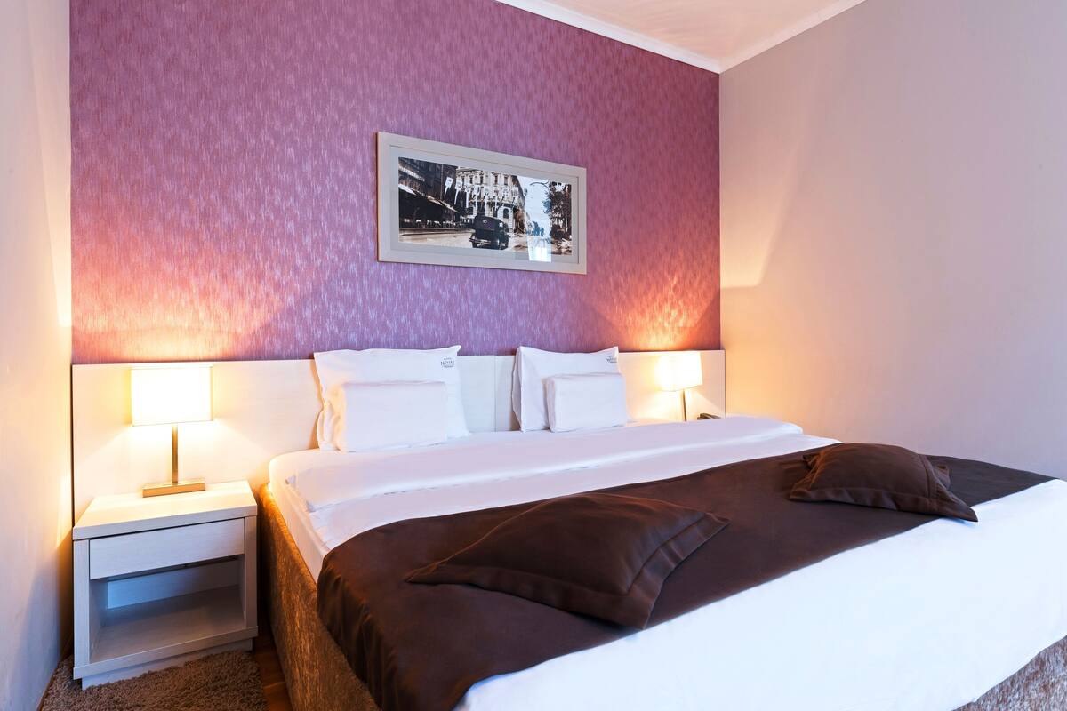 Habitación de hotel con una cama grande, dos mesitas de noche con lámparas, un cuadro enmarcado en la pared y una pared texturizada de color rosa.