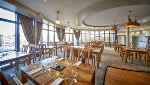 A spacious restaurant with multiple dining tables and chairs, large windows with curtains, and hanging pendant lights. - Lazaat Hotel (Cottingham)