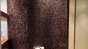 A bathroom with a curved shower wall tiled in a mosaic pattern, a toilet, and a towel rack with two rolls of toilet paper.