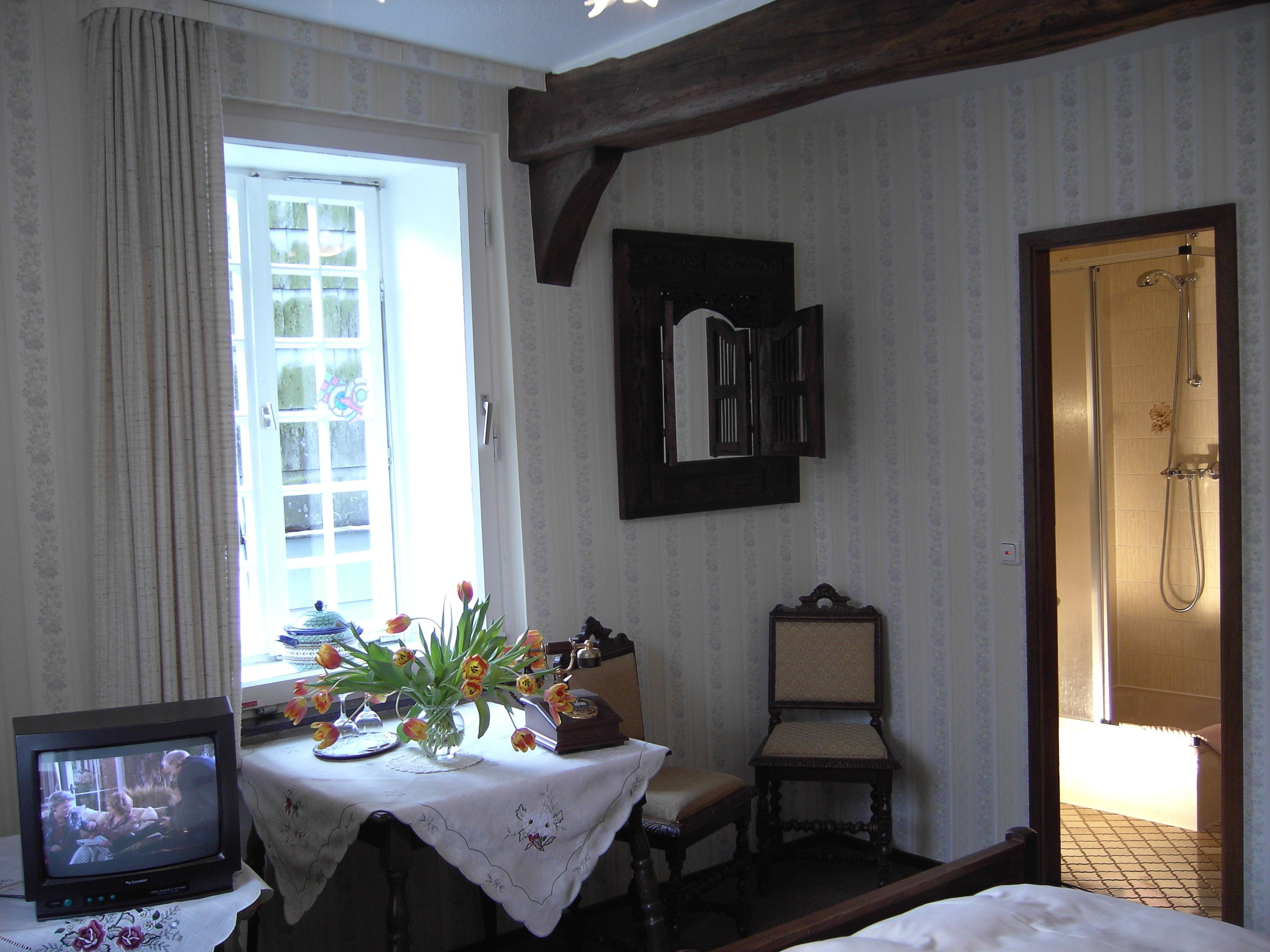 Quarto com cama, mesa com um vaso de flores, cadeira, televisão e porta que dá para um banheiro.